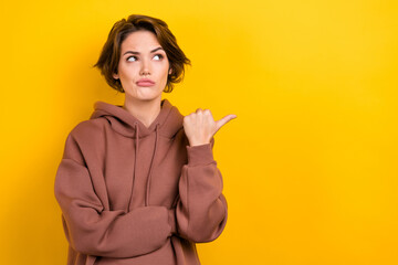 Photo of brunette hair girl indicate thumb finger empty space distrustful face wear brown hood shirt isolated yellow color background