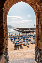 Archway through an ancient stone building leading to a beach with unrecognizable people in Cefalu,...