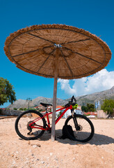 A serene beach scene in Greece, with a bike parked under a bamboo umbrella. It exudes relaxation and coastal charm, inviting viewers to envision themselves enjoying the sun and sea.