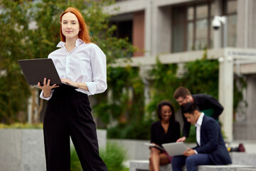 Fototapeta premium Portrait of serious, concentrated young woman, employee standing with laptop on street. Blurred employees on background