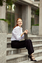 Fototapeta na wymiar Motivated and ambitious young woman, office manager sitting on stairs outdoors and working on laptop. Deadlines