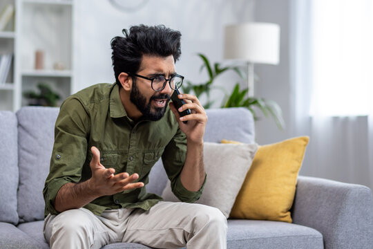 Angry and nervous man talking on the phone sitting on the sofa in the living room, hispanic man yelling at the interlocutor on the smartphone, dissatisfied with the service, the customer complains.