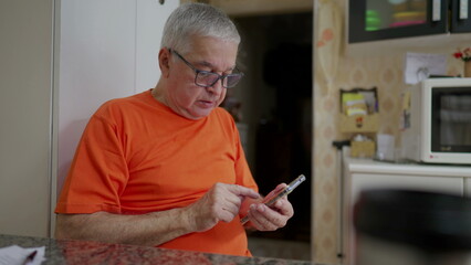 Senior man typing message on phone while sitting in kitchen. Older male person in 70s using modern technology