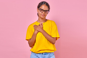 Young kind Indian woman presses palms to chest making gesture calling for active citizenship and helping charitable organizations stands on plain pink background. Caring, volunteering, activism
