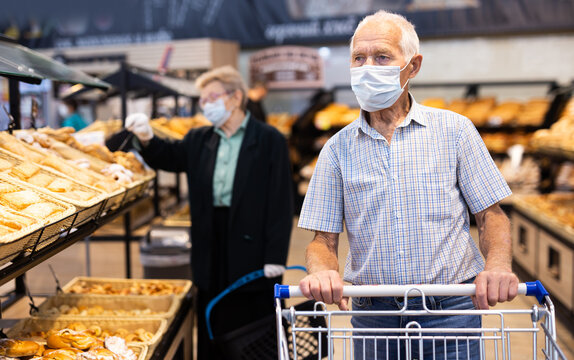 mature european man wearing mask and gloves with covid protection chooses buns and bread in supermarket bakery