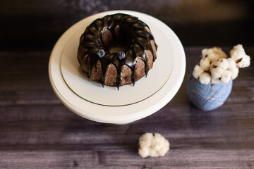 chocolate cupcake with icing on a white plate next to a vase with a cotton flower, on a dark background