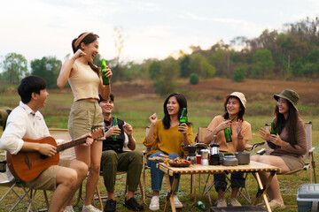 Group of Asian young adult people enjoy drinking a beer together during camping at the park. Asian young women and men celebrating with a bottle of a beer together.