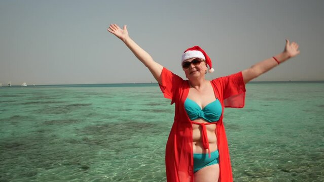 Funny senior woman in santa claus hats and sunglasses wearing in swimsuit on the sea beach, women dancing and wishes a merry Christmas and happy new year