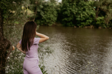 a girl in a lilac suit stands on the shore of a pond in summer. a walk in the park.