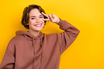 Portrait of young girl showing v sign near eye saying hi friends wear brown hoodie shirt isolated...