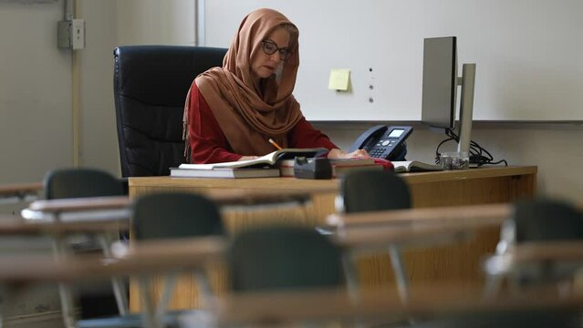 Portrait of happy smiling woman teacher wearing headscarf in empty school classroom working on assignments for students.