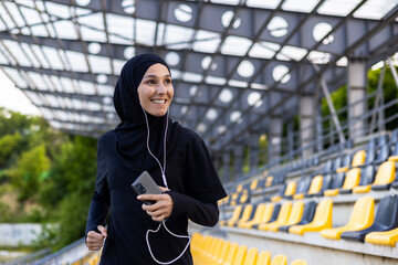 Young beautiful Muslim woman running in hijab in stadium, sportswoman smiling in headphones holding phone, listening to audio book.