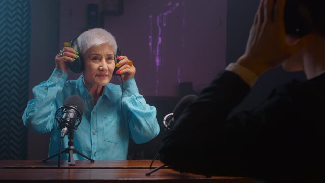 Serious old mature blogger woman in wireless headphones recordings podcast in a professional recording studio. An elderly lady on a radio interview. DJ creates live streaming on the Internet