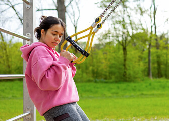 young woman in outside outdoor gym using cable chest for workout or parallel bars.girl female is resting sitting down drinking water from plastic bottle.pink hoodie park environment