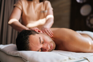Closeup face of relaxed young man getting back massage in spa salon lying on massage table closed eyes. Happy male client lying on stomach having full body treatment from professional masseuse.