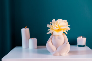 Close up white table with ceramic female body-shaped vase with rose flower, burning candles on turquoise blue wall background. Zen, cozy, meditation female bedroom decor. Self recovery place at home.