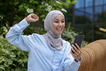Successful joyful muslim woman in hijab received online win notification, Dinka reading news and holding hand up triumph gesture celebrating sitting on bench outside office building in park