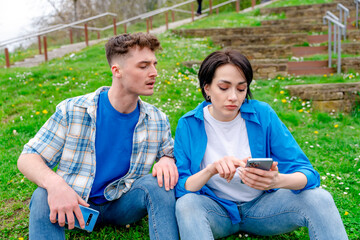 Sad couple using phones outdoors.  A man peeping into a woman's phone. Social media divide people concept.