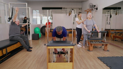 Group of elderly people doing Pilates Sessions with the help of female coach instructor. Older men and women using machines to stretch body and exercise, old age workout