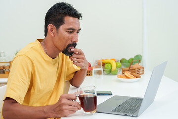 Bearded Asian man sitting in kitchen drinking morning coffee and looking at laptop computer. check stock trading on the stock market website to prepare for trading in the morning