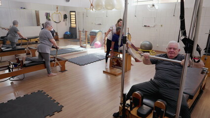 Fototapeta na wymiar Elderly people exercising with Pilates Machines in Group Session. Female Coach Instructor guiding seniors to move and stretch body. Old age workout routine
