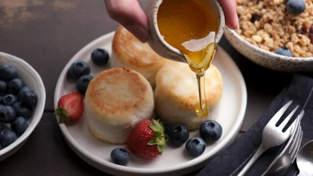 Slow motion pouring honey on syrniki, cottage cheese fritters served with fresh berries. Sweet breakfast or lunch meal