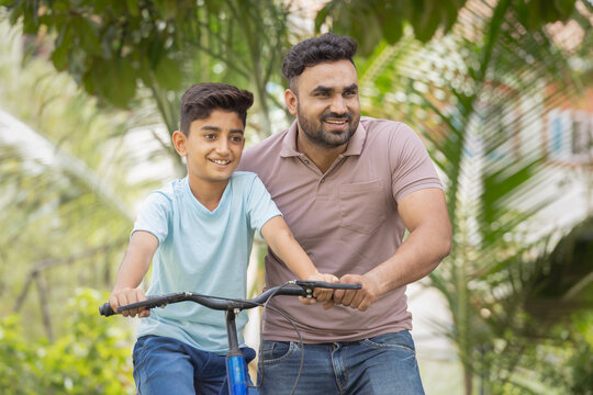 happy indian father helping his son to ride bicycle at park - concept of Childhood Milestones, Parental Guidance and memories - Powered by Adobe