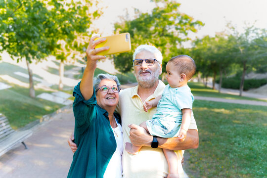 Happy grandparents couple with their baby grandchild in their arms taking a self-portrait with their cell phone