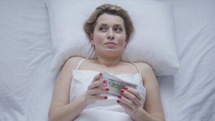 Cunning woman lying in bed and counting one hundred dollar bills and hiding money under the pillow, illegal earnings