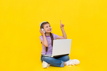 A child with a laptop. A little girl sits on the floor and communicates online on a laptop, wearing headphones. Internet communication for children. Yellow isolated background. copy space.