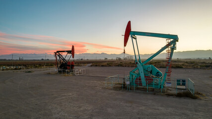 Pumpjack and transmission towers at sunset symbolizing energy transition. A pump jack pumping oil out of a well with silhouettes of electricity pylons and power line against a red sky.