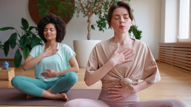 Female brunette trainer meditating with african woman and breathing on yoga mat