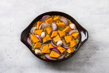 Lentils with pumpkin, red onion, garlic and spices, drenched in broth before baking on a gray textured table, top view. Cooking a delicious healthy vegan meal