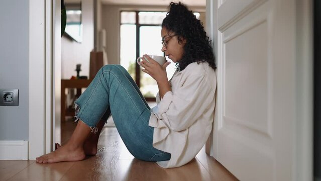 Happy curly african woman in eyeglasses drinking coffee on the floor at home