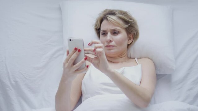 Top view woman scrolling web-pages on smartphone lying in bed late at night, watching posts in social networks and reading news and gossips, evening rituals