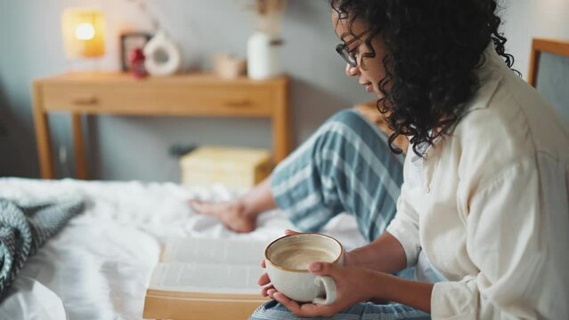 Pretty curly african woman in eyeglasses reading book with cup of coffee on the bed at home