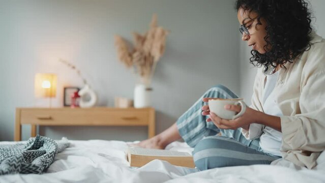 Relaxed curly african woman in eyeglasses reading book with cup of tea on the bed at home