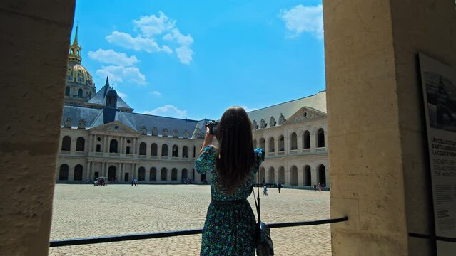 A young woman visiting and taking pictures of The Court of Honor of Hotel National des Invalides in Paris. A tourist looking at the Golden Dome of the Hotel des Invalides and rooftops of Paris.