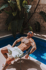 Chico joven musculado y tatuado posando  sin camiseta en piscina de estilo  boho en un hotel un dia...