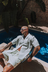 Chico joven tatuado posando con ropa de verano actual  en piscina estilo boho en un hotel un día...