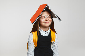 Funny little girl in school uniform and eyeglasses with book on head and dreaming with closed eyes,...