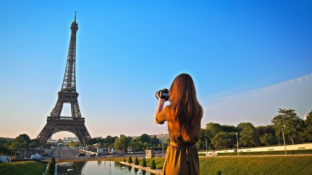 A beautiful woman in a dress uses a camera to picture the romantic city of Paris, France. Female tourist taking pictures of the historical Tower and enjoying the view of a golden sunset.