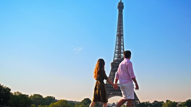 A young couple enjoys the view of Paris pointing at the Tower. A romantic woman and her boyfriend walk together holding hands at golden sunset in Paris, France.