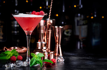 Clover club alcoholic cocktail drink with dry gin, raspberry syrup, lime juice, quail egg white and ice in glass. Black bar counter background, bar tools and bottles