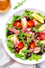 Nicoise salad with canned tuna, cherry tomatoes, boiled eggs, green beans and red olives on plate, white table background, top view