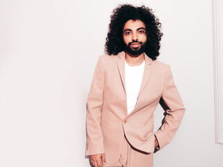Handsome  hipster  model. Unshaven Arabian man dressed in elegant beige suit . Fashion male with long curly hairstyle posing near grey wall in studio. Isolated