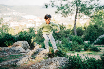 Naklejka na ściany i meble School kid child in vibrant clothes jumping over granite rock boulders in the city park with mountain forest view in the background. Boy leaping across rocks in the park.