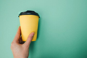 Yellow paper cup with coffee in a female hand on a green background. Front view