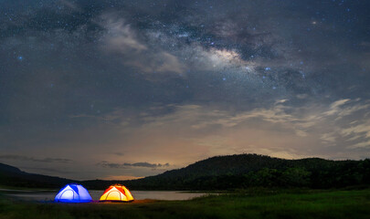 Fototapeta na wymiar Panorama of the night sky with camping tents and lakes, surrounded by mountains. Milky way and stars on dark background with noise and grain. Long exposure shot with white balance selected.