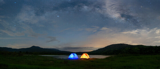 Panorama of the night sky with camping tents and lakes, surrounded by mountains. Milky way and...